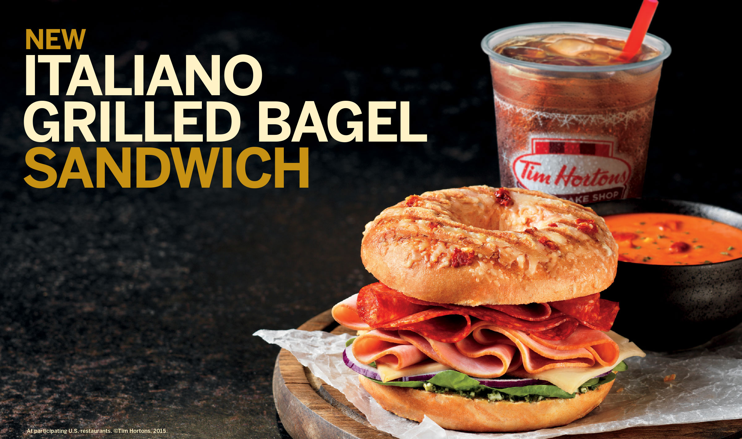 Ad for an Italian bagel sandwich, with the sandwich, a bowl of tomato soup, and a cup of iced tea sitting on a cutting board against a dark background